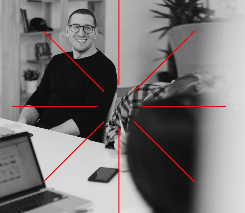 Black and white image of a man smiling with a computer. A red-line graphic element in center that creates a star.